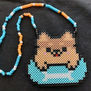 May include: A beaded necklace with a brown and tan perler bead design of a cartoon animal with a blue and white perler bead design of a boat. The necklace is made with blue, white, orange, and black beads.