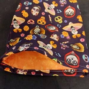 May include: A dark blue fabric pouch with a repeating pattern of sugar skulls, sombreros, musical notes, and flowers. The interior is a solid orange color. The pouch is open, revealing the orange lining.