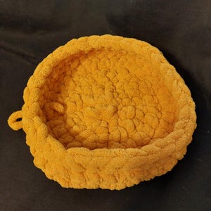 May include: A round, mustard-yellow crochet pet bed. The bed has a thick, textured appearance and a low, raised edge. A small loop is attached to the side. The bed is designed for small pets.
