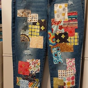 May include: Blue denim jeans with colorful patchwork details. The patches feature various patterns and colors, including floral, geometric, and abstract designs. The jeans have a distressed look with frayed hems.