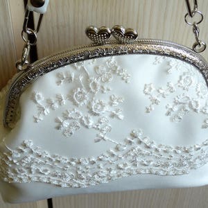 May include: A white satin clutch bag with a silver metal frame and a delicate floral lace design. The bag has a chain strap and a heart-shaped clasp.