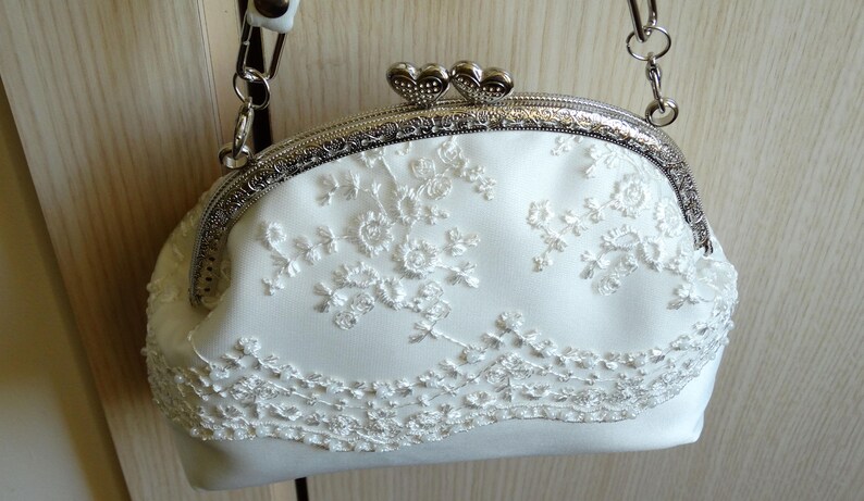 May include: A white satin clutch bag with a silver metal frame and a delicate floral lace design. The bag has a chain strap and a heart-shaped clasp.