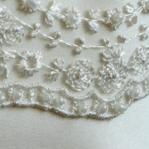 May include: White satin fabric with a delicate lace trim featuring a floral design and pearl accents.
