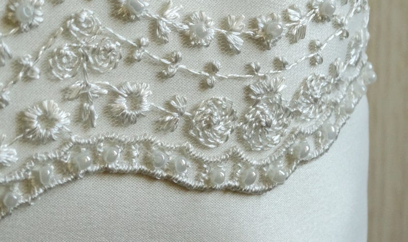 May include: White satin fabric with a delicate lace trim featuring a floral design and pearl accents.