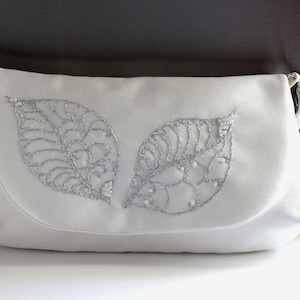 May include: White clutch bag with a flap closure. The bag features two large, silver embroidered leaf designs on the front. The silver embroidery is detailed with sequins. The bag has a silver chain strap.