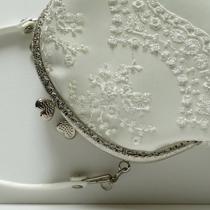 May include: A white satin clutch purse with a silver metal frame and a delicate lace design. The purse has a silver chain strap and a heart-shaped clasp.