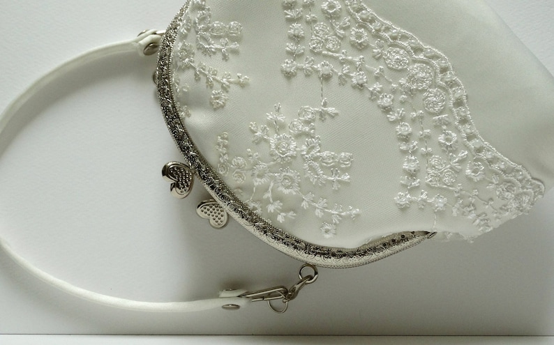 May include: A white satin clutch purse with a silver metal frame and a delicate lace design. The purse has a silver chain strap and a heart-shaped clasp.