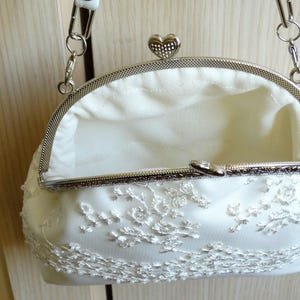 May include: A white satin clutch bag with a silver chain strap and a heart-shaped clasp. The bag is decorated with white lace.