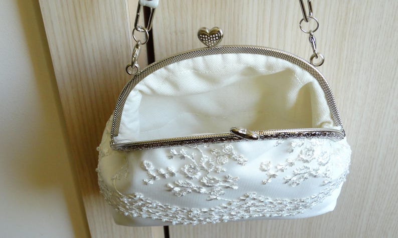May include: A white satin clutch bag with a silver chain strap and a heart-shaped clasp. The bag is decorated with white lace.