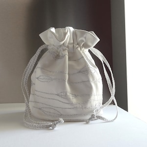 Könnte beinhalten: Eine weiße Kordeltasche mit silbernen Stickereien von Blättern. Die Tasche ist aus einem weichen, seidenartigen Stoff gefertigt und hat ein schlichtes, elegantes Design.