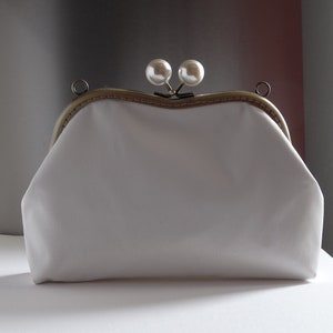 Pode incluir: Uma bolsa de m&atilde;o branca de tecido com uma moldura de metal dourado e dois detalhes de p&eacute;rolas no fecho.