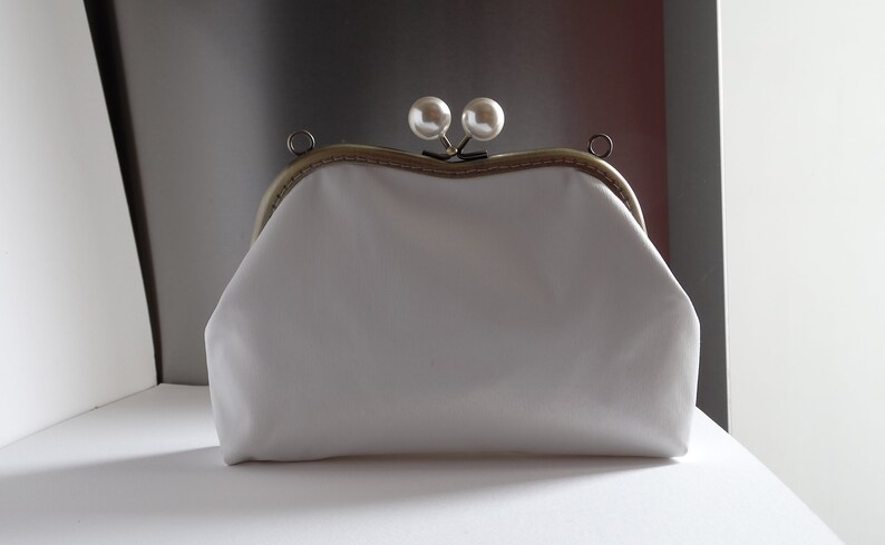 Pode incluir: Uma bolsa de m&atilde;o branca de tecido com uma moldura de metal dourado e dois detalhes de p&eacute;rolas no fecho.