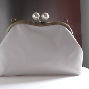 Pode incluir: Uma bolsa de m&atilde;o branca de tecido com uma moldura de metal dourado e dois enfeites de p&eacute;rolas.