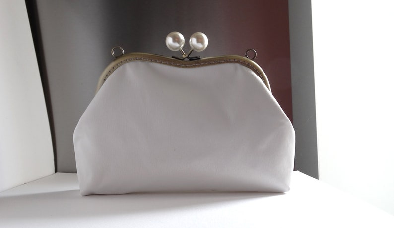 Pode incluir: Uma bolsa de m&atilde;o branca de tecido com uma moldura de metal dourado e dois enfeites de p&eacute;rolas.
