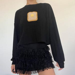May include: Black long-sleeve sweater with a square, crocheted patch of toast and butter. The sweater is paired with a black, tiered lace skirt. The outfit is casual and stylish.