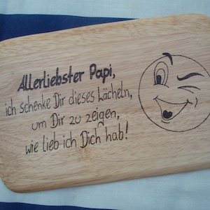 Può includere: Un tagliere di legno con un messaggio inciso che recita "Allerliebster Papi, ich schenke Dir dieses Lächeln, um Dir zu zeigen, wie lieb ich Dich hab!" e un'emoticon sorridente con un occhiolino.