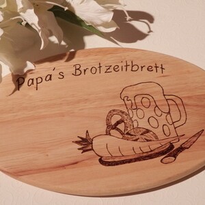 Puede incluir: Una tabla de cortar ovalada de madera con el texto "Papa's Brotzeitbrett" grabado en ella. La tabla presenta un diseño de jarra de cerveza, pretzel y una zanahoria.