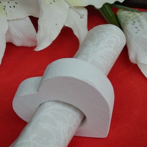 Op de afbeelding: Een witte houten hartvormige servetring met een witte, gepatenteerde stoffen ring erin. De ring ligt op een rood oppervlak.