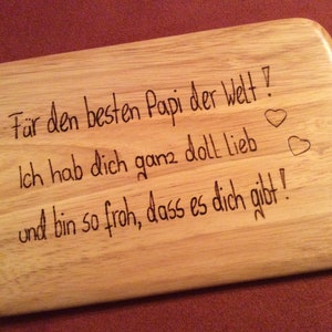 Può includere: Un tagliere di legno con un messaggio inciso. Il messaggio recita: "Per il papà migliore del mondo! Ti voglio molto bene e sono così felice che tu sia qui!" Ci sono due cuori disegnati alla fine del messaggio.