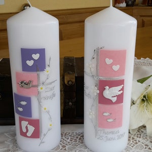 Puede incluir: Dos velas de bautismo blancas con cuadrados rosas y morados, palomas plateadas y peces plateados. El texto "Zur Taufe" y "Theresa 25. Juni 2017" está escrito en las velas.