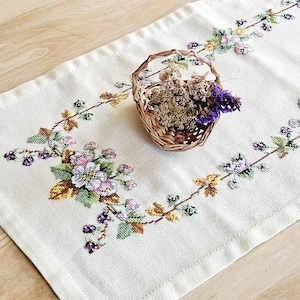 May include: Cream-colored embroidered table runner with floral and leaf patterns in pink, purple, green, and yellow. A small woven basket filled with dried flowers sits on the runner.