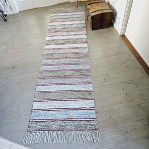 Puede incluir: Una alfombra larga y estrecha, tejida, con fondo blanco y rayas rojas, grises y azules. La alfombra tiene flecos en ambos extremos.