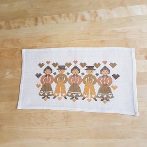 Puede incluir: Un camino de mesa de lino blanco con un diseño de punto de cruz de cinco figuras de la mano, en marrón, naranja y amarillo. Las figuras están rodeadas de corazones marrones y blancos.