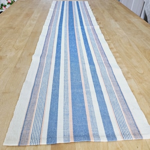May include: A white table runner with blue and pink stripes. The runner is made of a woven fabric and has a slightly textured surface.