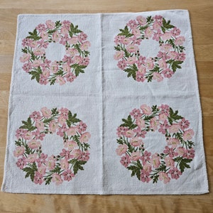 May include: White linen tablecloth with four pink floral wreaths printed on it. The wreaths are made of pink flowers with green leaves.