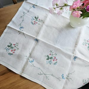 Op de afbeelding: Een wit linnen tafelkleed met delicate handgeborduurde motieven van rode aardbeien, groene bladeren en kleine blauwe vlinders. Het borduurwerk is in een verspreid patroon gerangschikt over de gevouwen en getextureerde stof.