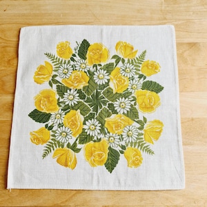 Könnte beinhalten: Ein quadratisches, cremefarbenes Küchentuch mit Blumenmuster. Das Design zeigt einen zentralen Strauß aus gelben Rosen, weißen Gänseblümchen und grünen Blättern. Die Blumen sind kreisförmig angeordnet und erzeugen eine lebendige und fröhliche Ästhetik. Das Tuch besteht aus einem gewebten Stoff.