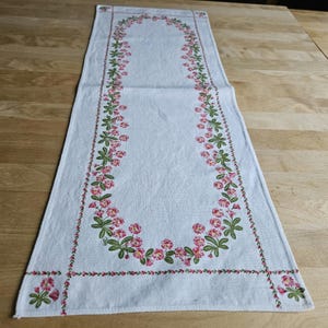 May include: White table runner with a floral design featuring pink flowers and green leaves. The flowers are arranged in a border around the edge of the runner.