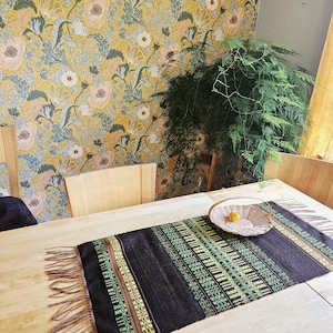 May include: A dining table with a black and green patterned table runner with fringe. A woven basket with a yellow object sits on the table. The background features floral wallpaper and a large green plant.