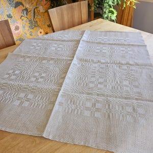 Könnte beinhalten: Eine beige Tischdecke aus Leinen mit einem dezenten geometrischen Muster. Die Tischdecke ist in der Mitte gefaltet und auf einem Holztisch ausgelegt.