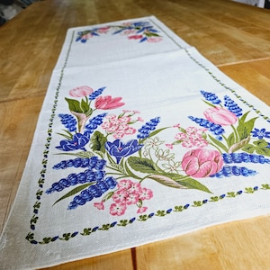May include: White table runner with a floral design featuring blue, pink, and green flowers. The design is centered on the runner and has a border of green leaves.