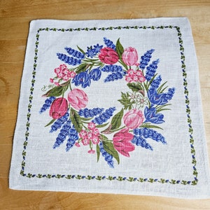 May include: A white linen tea towel with a green border and a floral design. The design features pink and blue flowers, and green leaves.