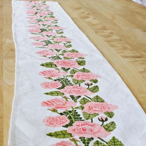 May include: A white table runner with a pink and green floral design. The design features a row of pink roses with green leaves.