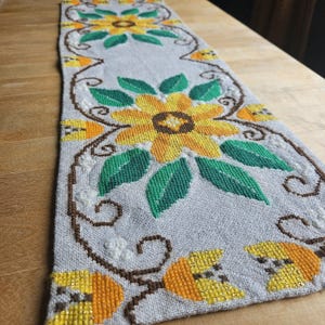 May include: A white linen table runner with embroidered yellow sunflowers and green leaves. The runner has a brown border and a scalloped edge.