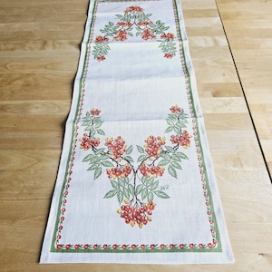 May include: A white linen table runner with a green border and a floral design featuring red berries and green leaves. The runner has the text "Chisa R" printed on it.