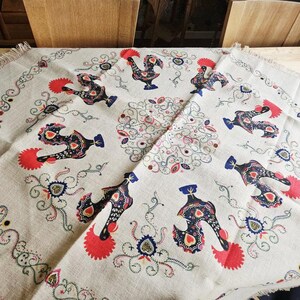 May include: A beige linen tablecloth with a repeating pattern of stylized roosters in red, blue, and black. The design includes floral and heart motifs, with a decorative border. The tablecloth is folded, showing the pattern's continuity.