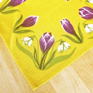 May include: A yellow fabric placemat with a floral pattern featuring purple and white crocuses. The flowers are arranged in a circular pattern around the edge of the placemat.