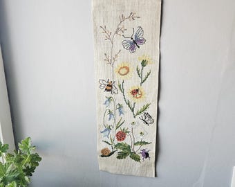 Puede incluir: Un tapiz de pared bordado a punto de cruz con un diseño floral e insectos. La tela beige muestra una mariposa, una abeja, un caracol y varias flores en tonos amarillos, azules y morados. El tapiz está suspendido de una barra de metal decorativa.