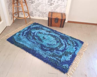Puede incluir: Una alfombra rectangular con un patrón arremolinado en tonos azules y turquesas. La alfombra tiene una textura peluda y flecos beige en los extremos cortos. La alfombra está colocada sobre un suelo de madera clara.
