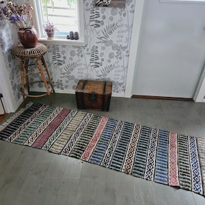 Puede incluir: Una alfombra rayada colorida con un patrón geométrico en tonos de azul, verde, rojo y blanco. La alfombra está colocada sobre un suelo de madera delante de un cofre de madera.