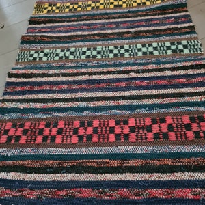 Puede incluir: Una alfombra rectangular tejida multicolor con rayas horizontales y patrones de cuadros. La alfombra presenta una variedad de colores, incluyendo azul, rojo, marrón, verde y amarillo. La alfombra está hecha de un material texturizado.