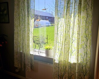 May include: White sheer curtains with a green leaf pattern hang in a window. The curtains are open, revealing a view of a green lawn and a building in the distance.