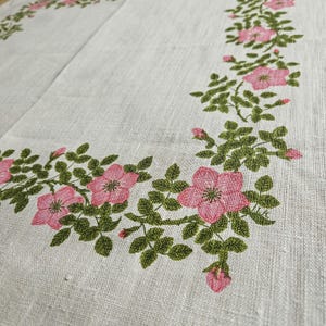 May include: A white linen tablecloth with a pink and green floral border. The border features a repeating pattern of pink roses and green leaves.