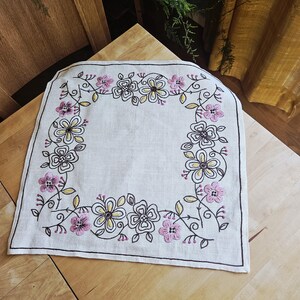 Puede incluir: Un mantel individual cuadrado de lino blanquecino con un diseño de bordado floral. El bordado presenta flores rosas y amarillas con contornos negros, dispuestas en un borde. Colocado sobre una superficie de madera clara.