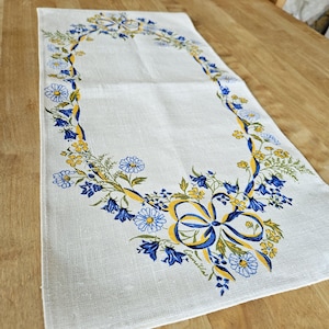 May include: A white linen table runner with a blue and yellow floral border. The border features a repeating pattern of bluebells, daisies, and yellow ribbons. The text "Linella" is embroidered in the lower right corner.