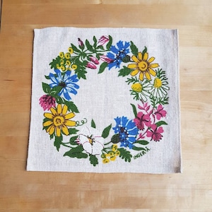 Könnte beinhalten: Eine weiße Leinenserviette mit einem farbenfrohen Blumengirlanden-Design. Die Girlande besteht aus gelben, blauen, rosa und weißen Blumen. Der Text "INGERIA" ist in der rechten unteren Ecke gedruckt.